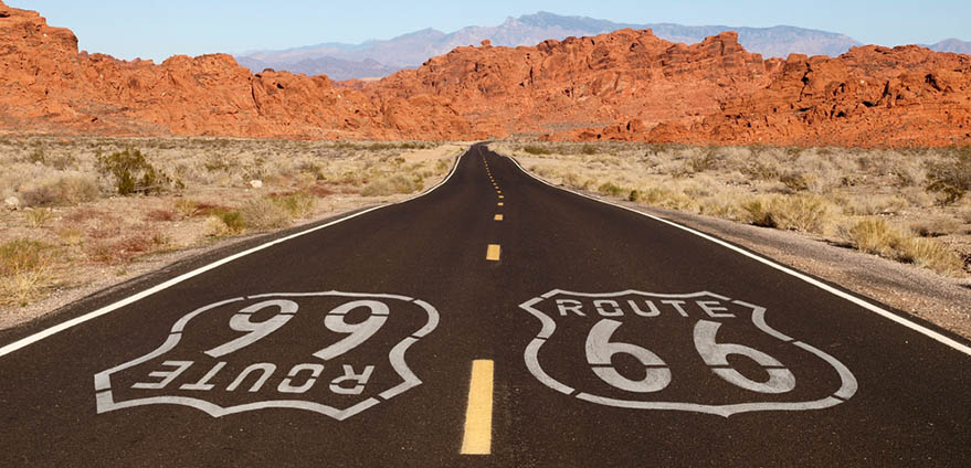 Route 66 is written on a highway with the Arizona desert and red mountains in the background against a clear blue sky